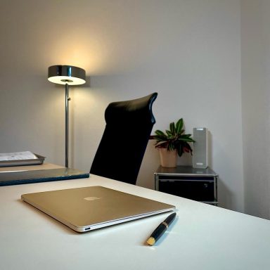 Schreibtisch mit Laptop, Stuhl, Tischlampe und Pflanze im Hintergrund.