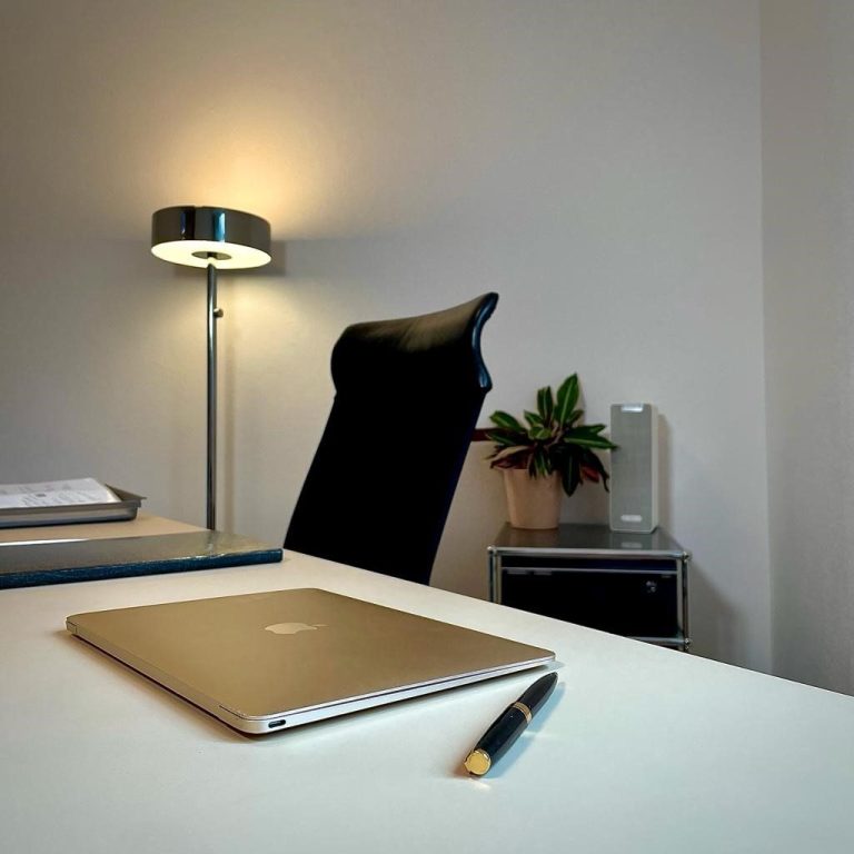 Ein moderner Schreibtisch mit Laptop, Stuhl, Lampe und einer Pflanze in einem hellen Raum.