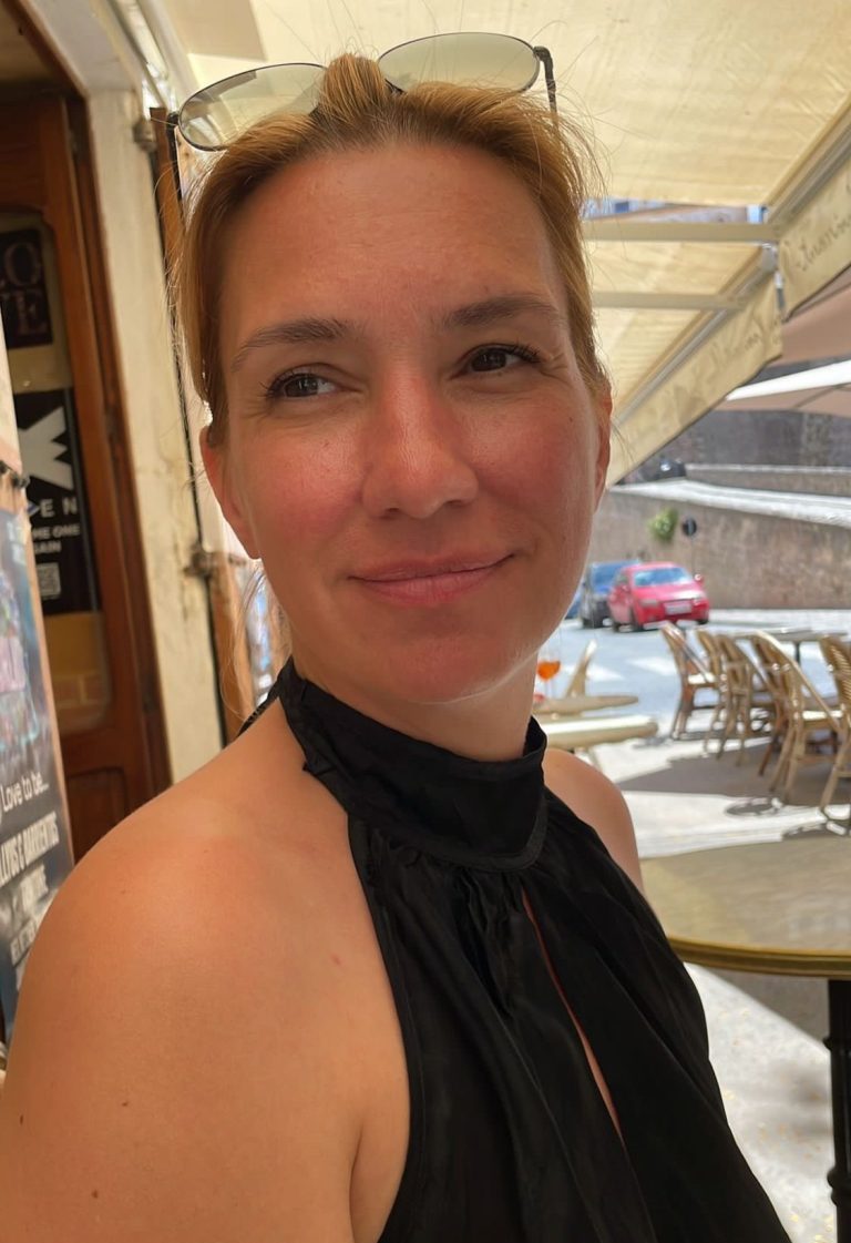 Frau mit blonden Haaren, lächelnd, in einem Café mit Sonnenschein im Hintergrund.