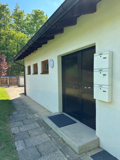 Haus mit schwarzer Eingangstür, Briefkästen rechts, gepflastertem Weg und Bäumen im Hintergrund.
